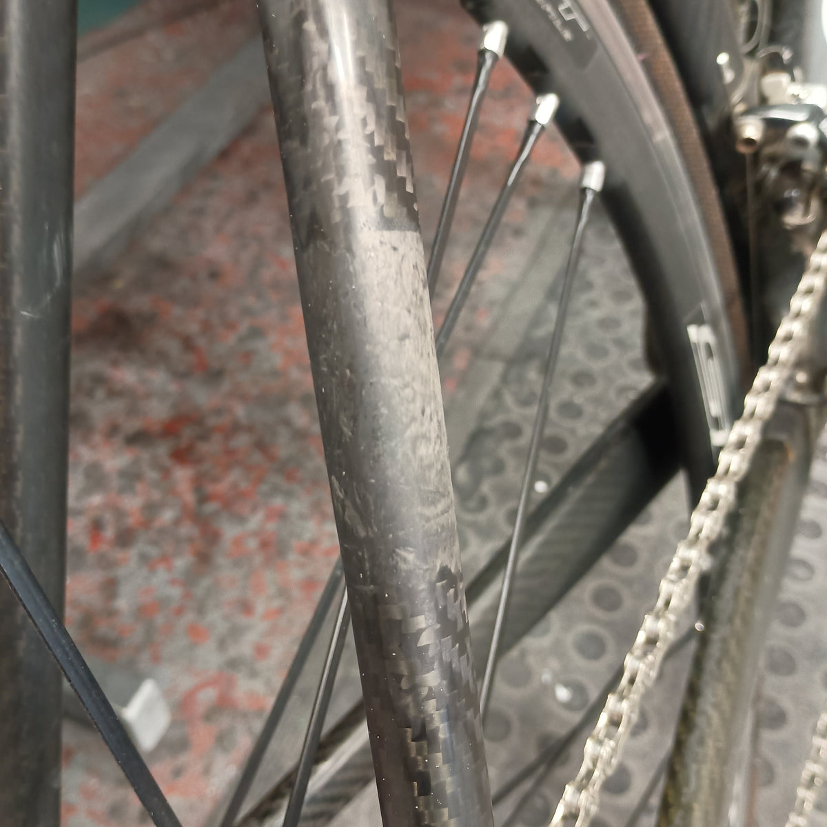 Close-up of a carbon fiber bicycle frame and wheel with visible scuffs and wear marks on the tube, alongside a chainring and chain.
