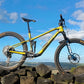 A yellow and black full-suspension mountain bike is photographed outdoors on a stone wall under a bright blue sky with white clouds.