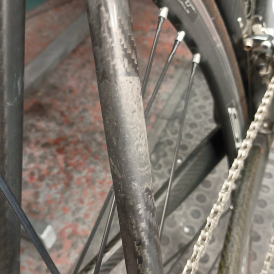 Close-up of a carbon fiber bicycle frame and wheel with visible scuffs and wear marks on the tube, alongside a chainring and chain.